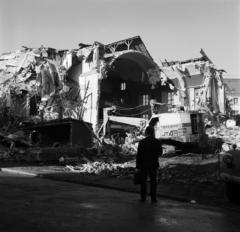 Sama työmaa romahduksen jälkeen, talon saliosan holvirakenne on vaativa purettava ja jos tämä Lokomo Teräsmies 43 kuokkakaivulaitteella oli työmaan ainoa kaivinkone, on näin korkean rakennuksen purkaminen vaikeaa, koneessa on vasen liuku-ovi pudonnut paikoiltaan ja nojaa koneistoon, molempien rumpujen vaijerit roikkuvat löysinä, joten kuljettaja on poistunut koneesta kiinnittämättä jarruja, kuva Hakli Kari, Helsingin kaupunginmuseo.