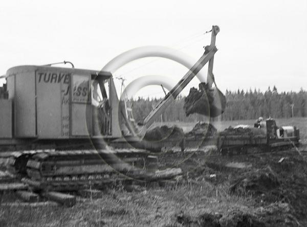 Teksti Luston kuvatiedoissa: Turvetta nostetaan junanvaunun Åkerman 300 (Turve-Jussi) -kaivinkoneella Seppälän suolla. ""Turve Jussin" koura nostaa suosta kokonaisen kuutiometrin suota rautatievaunuun." (alkuperäinen teksti kuvan takana). Vesterinen, Emil 12. - 13.7.1943.