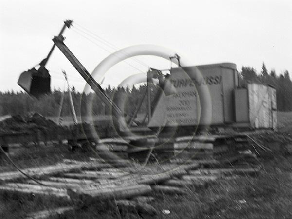 Teksti Luston kuvatiedoissa: Turvetta nostetaan junanvaunun Åkerman 300 (Turve-Jussi) -kaivinkoneella Seppälän suolla. ""Turve Jussin" koura nostaa suosta kokonaisen kuutiometrin suota rautatievaunuun." (alkuperäinen teksti kuvan takana). Vesterinen, Emil 12. - 13.7.1943.