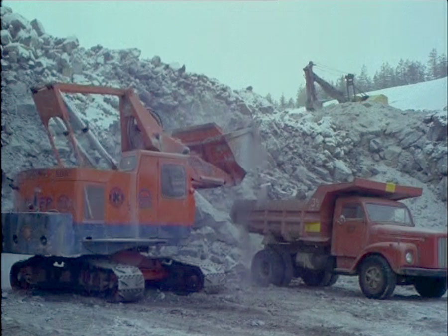 Filmissä näkyy erikoinen kaivinkone, joka on amerikkalainen Koehring Skooper. Kuva on epäselvä, mutta näyttää siltä, että koneessa on Vesto Oy:n merkki kyljessä. Skooperin puomin järjestely oli melko erikoinen, mutta siinä on selvästi järkeä, kun ajattelee lastauskoneelta vaadittavia ominaisuuksia. Kauhan eteen-taakse -liike onnistui paremmin kuin tavallisessa vaijereilla tehdyssä pistokaivinkoneessa. Sylinterejä (sylinteripareja) ei tarvita tässä järjestelyssä yhtään enempää kuin tavallisessa hydraulisessa kuokkakaivinkoneessa. Kolme riittää. Raskaimmat liikkeet on onnistuttu järjestämään niin, että sylinterit työskentelevät ulossuuntaan. Siksi ylävaunun päällä olevat sylinterit on käännetty taaksepäimn ja yhdistetty puomiin henkseleiden välityksellä.