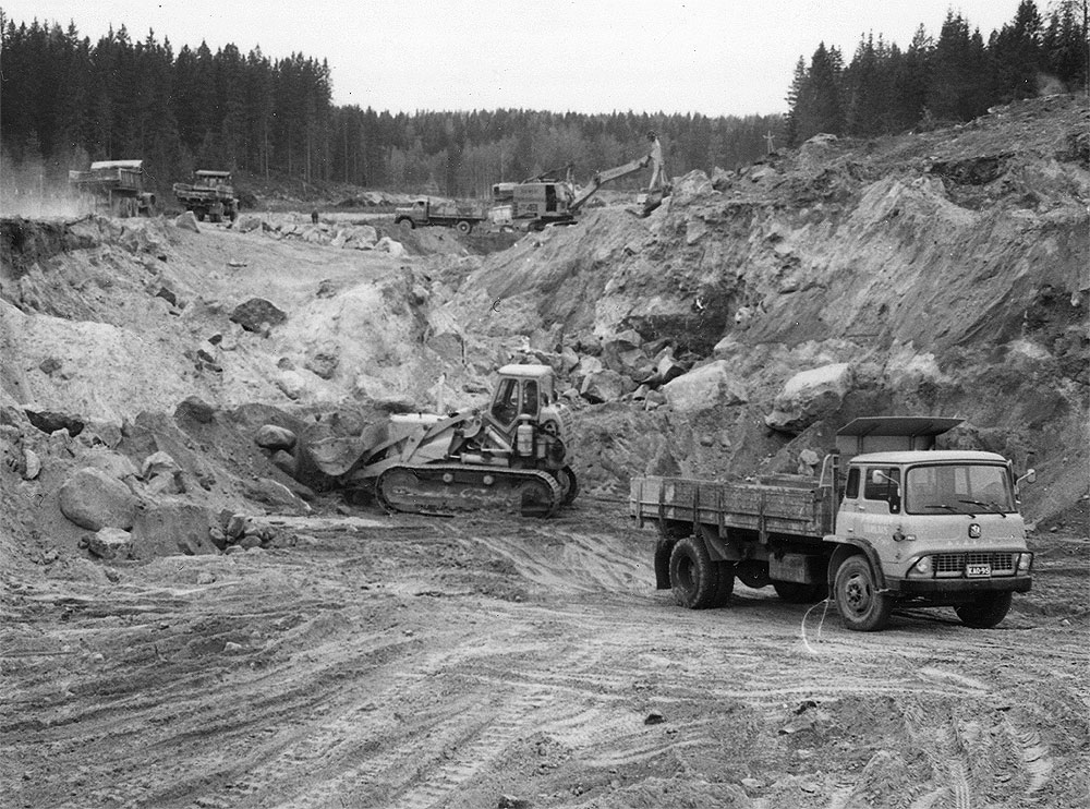 Kuvassa näkyvät autot: Kraz, tuntematon, Magirus-Deutz 150D (?), Bedford KHTC 14 (?), kavinkoneet Lokomo Teräsmies JT22, Lokomo Teräsmies JT43 ja Caterpillar Traxcavator 977, kanavan kaivamista Lauritsalassaa, Liikennevirasto